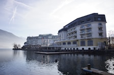 A large, elegant hotel building with multiple floors and balconies is situated alongside a calm body of water, possibly a lake. The scene is set in a mountainous area with misty hills in the background and a clear sky above. The architecture is traditional with a combination of white and beige colors, and there are small decorative plants on the balconies. The water reflects the building and surrounding landscape, adding to the tranquil atmosphere.