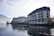 A large, elegant hotel building with multiple floors and balconies is situated alongside a calm body of water, possibly a lake. The scene is set in a mountainous area with misty hills in the background and a clear sky above. The architecture is traditional with a combination of white and beige colors, and there are small decorative plants on the balconies. The water reflects the building and surrounding landscape, adding to the tranquil atmosphere.
