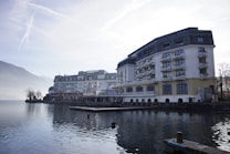 A large, elegant hotel building with multiple floors and balconies is situated alongside a calm body of water, possibly a lake. The scene is set in a mountainous area with misty hills in the background and a clear sky above. The architecture is traditional with a combination of white and beige colors, and there are small decorative plants on the balconies. The water reflects the building and surrounding landscape, adding to the tranquil atmosphere.