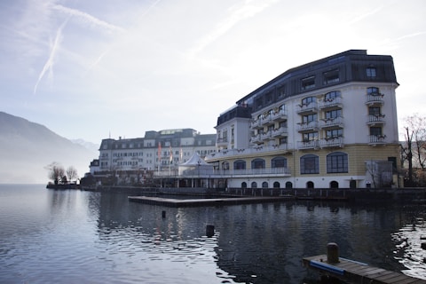 A large, elegant hotel building with multiple floors and balconies is situated alongside a calm body of water, possibly a lake. The scene is set in a mountainous area with misty hills in the background and a clear sky above. The architecture is traditional with a combination of white and beige colors, and there are small decorative plants on the balconies. The water reflects the building and surrounding landscape, adding to the tranquil atmosphere.
