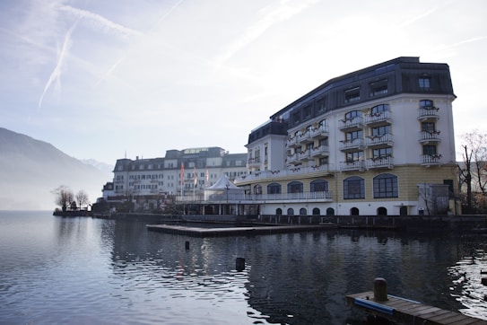 A large, elegant hotel building with multiple floors and balconies is situated alongside a calm body of water, possibly a lake. The scene is set in a mountainous area with misty hills in the background and a clear sky above. The architecture is traditional with a combination of white and beige colors, and there are small decorative plants on the balconies. The water reflects the building and surrounding landscape, adding to the tranquil atmosphere.