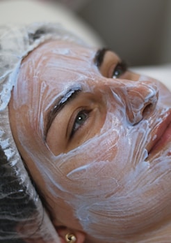 A person lies down with a creamy facial mask applied evenly over the face. The individual wears a protective cap, and the eyes appear relaxed yet focused. The texture of the cream is smooth and shiny.