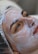 A person lies down with a creamy facial mask applied evenly over the face. The individual wears a protective cap, and the eyes appear relaxed yet focused. The texture of the cream is smooth and shiny.