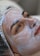 A person lies down with a creamy facial mask applied evenly over the face. The individual wears a protective cap, and the eyes appear relaxed yet focused. The texture of the cream is smooth and shiny.