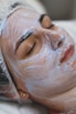 A person is lying down with their eyes closed, seemingly relaxed, while a thick layer of a white facial cream or mask is applied on their face. The head is covered with a protective hairnet, and the person's features indicate they are receiving a beauty treatment.
