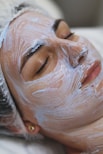 A person is lying down with their eyes closed, seemingly relaxed, while a thick layer of a white facial cream or mask is applied on their face. The head is covered with a protective hairnet, and the person's features indicate they are receiving a beauty treatment.