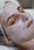 A person is lying down with their eyes closed, seemingly relaxed, while a thick layer of a white facial cream or mask is applied on their face. The head is covered with a protective hairnet, and the person's features indicate they are receiving a beauty treatment.