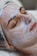 A person is lying down with their eyes closed, seemingly relaxed, while a thick layer of a white facial cream or mask is applied on their face. The head is covered with a protective hairnet, and the person's features indicate they are receiving a beauty treatment.