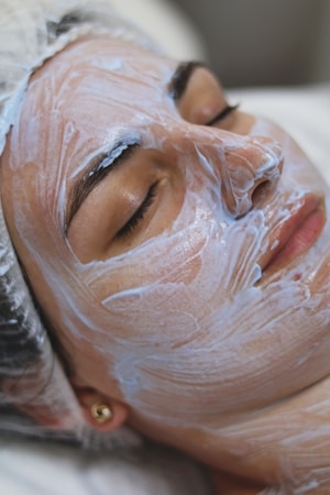 A person is lying down with their eyes closed, seemingly relaxed, while a thick layer of a white facial cream or mask is applied on their face. The head is covered with a protective hairnet, and the person's features indicate they are receiving a beauty treatment.