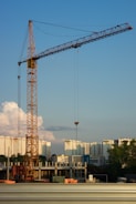 A construction site with a large crane towering over partially built structures. In the background, high-rise apartment buildings are visible under a clear blue sky with some scattered clouds.
