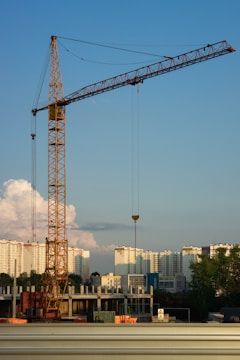 A construction site with a large crane towering over partially built structures. In the background, high-rise apartment buildings are visible under a clear blue sky with some scattered clouds.