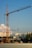 A construction site with a large crane towering over partially built structures. In the background, high-rise apartment buildings are visible under a clear blue sky with some scattered clouds.