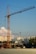 A construction site with a large crane towering over partially built structures. In the background, high-rise apartment buildings are visible under a clear blue sky with some scattered clouds.