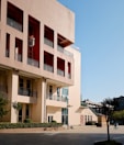 A modern urban building with a beige and red facade, featuring multiple rows of windows and balconies. A courtyard in front of the building contains neatly trimmed bushes and trees, along with several bicycles parked beside a path.