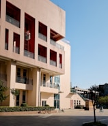 A modern urban building with a beige and red facade, featuring multiple rows of windows and balconies. A courtyard in front of the building contains neatly trimmed bushes and trees, along with several bicycles parked beside a path.