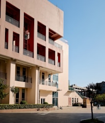 A modern urban building with a beige and red facade, featuring multiple rows of windows and balconies. A courtyard in front of the building contains neatly trimmed bushes and trees, along with several bicycles parked beside a path.