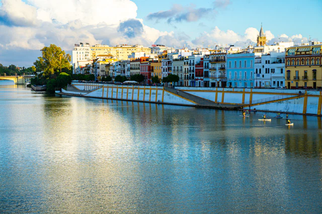 Golden-hour view across the Guadalquivir River toward Triana's colorful riverfront houses, with a bridge and church tower in the distance.