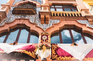 An ornate facade displaying intricate architectural details with decorative brickwork, scrolling blue patterns, and arched windows. The area below the windows is adorned with elaborate fabric banners, featuring red and gold colors and floral decorations. A distinctive feature in the center includes a sun-like emblem surrounded by flowers.
