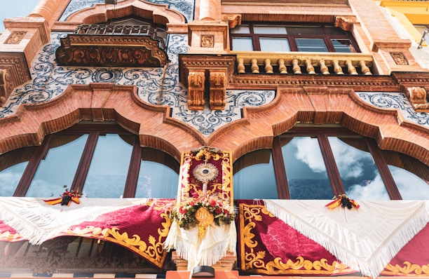 An ornate facade displaying intricate architectural details with decorative brickwork, scrolling blue patterns, and arched windows. The area below the windows is adorned with elaborate fabric banners, featuring red and gold colors and floral decorations. A distinctive feature in the center includes a sun-like emblem surrounded by flowers.