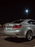 A silver Lexus sedan is parked in a dimly lit parking lot at night. The car's rear section is shown, highlighting polished metal rims and sharp red tail lights. The background includes a bright street lamp and distant, blurred lights possibly from a building or signs.
