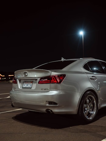 A silver Lexus sedan is parked in a dimly lit parking lot at night. The car's rear section is shown, highlighting polished metal rims and sharp red tail lights. The background includes a bright street lamp and distant, blurred lights possibly from a building or signs.