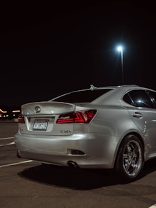 A silver Lexus sedan is parked in a dimly lit parking lot at night. The car's rear section is shown, highlighting polished metal rims and sharp red tail lights. The background includes a bright street lamp and distant, blurred lights possibly from a building or signs.