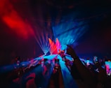 A live concert event with vibrant lighting effects. The stage is bathed in a mix of blue and red lights, creating a striking visual. The crowd in the foreground holds up smartphones, capturing the performance. The performer stands at the center of the stage, illuminated by spotlights.