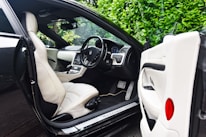 The image features the interior of a luxury car, showcasing a sleek design with cream leather seats. The steering wheel is detailed and the dashboard is modern with a digital display. The car door is open, revealing the spacious seating and finely crafted interior. Lush green foliage can be seen through the windows, adding a natural contrast to the luxury setting.