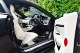 The image features the interior of a luxury car, showcasing a sleek design with cream leather seats. The steering wheel is detailed and the dashboard is modern with a digital display. The car door is open, revealing the spacious seating and finely crafted interior. Lush green foliage can be seen through the windows, adding a natural contrast to the luxury setting.