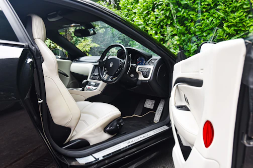 The image features the interior of a luxury car, showcasing a sleek design with cream leather seats. The steering wheel is detailed and the dashboard is modern with a digital display. The car door is open, revealing the spacious seating and finely crafted interior. Lush green foliage can be seen through the windows, adding a natural contrast to the luxury setting.