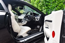 The image features the interior of a luxury car, showcasing a sleek design with cream leather seats. The steering wheel is detailed and the dashboard is modern with a digital display. The car door is open, revealing the spacious seating and finely crafted interior. Lush green foliage can be seen through the windows, adding a natural contrast to the luxury setting.