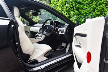The image features the interior of a luxury car, showcasing a sleek design with cream leather seats. The steering wheel is detailed and the dashboard is modern with a digital display. The car door is open, revealing the spacious seating and finely crafted interior. Lush green foliage can be seen through the windows, adding a natural contrast to the luxury setting.