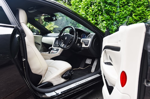 The image features the interior of a luxury car, showcasing a sleek design with cream leather seats. The steering wheel is detailed and the dashboard is modern with a digital display. The car door is open, revealing the spacious seating and finely crafted interior. Lush green foliage can be seen through the windows, adding a natural contrast to the luxury setting.