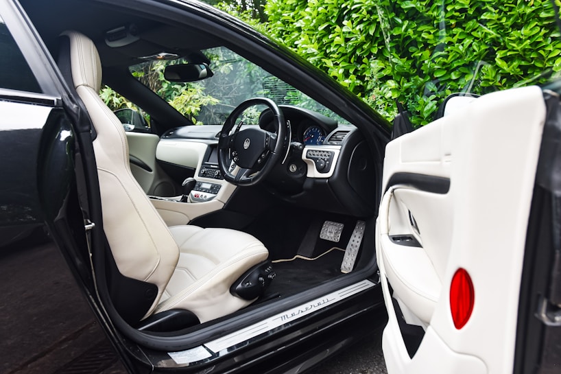 The image features the interior of a luxury car, showcasing a sleek design with cream leather seats. The steering wheel is detailed and the dashboard is modern with a digital display. The car door is open, revealing the spacious seating and finely crafted interior. Lush green foliage can be seen through the windows, adding a natural contrast to the luxury setting.