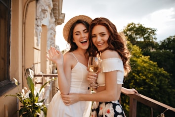 Two women are smiling on a balcony with lush greenery in the background. One woman is wearing a straw hat and white dress, while the other is holding a glass of wine and wearing an off-shoulder top with a floral skirt. Sunlight bathes the scene, creating a warm, inviting atmosphere.