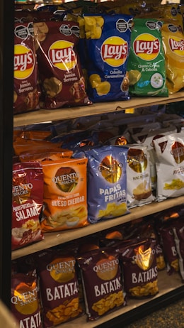 A supermarket shelf displaying a variety of snack chips. The shelf includes multiple brands such as Lay's and Quento Snacks, featuring flavors like Jamón Serrano, Cebolla Caramelizada, Queso Crema y Cebolla, Queso Cheddar, and Salame. The packaging is colorful, with dominant hues of red, blue, green, orange, and white.