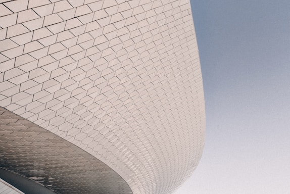 The image features a section of modern architecture with a curved, tiled facade. The tiles form an intricate pattern with geometric shapes, mostly diamond-like in appearance. The surface reflects subtle lighting, enhancing its texture and dimensionality against a soft blue sky.