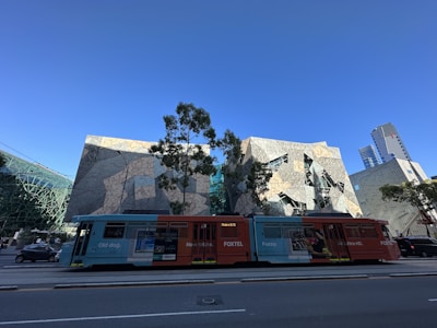 A modern tram in bright colors travels on a city street in the foreground. Behind it is a striking, geometrically designed building with a faceted exterior that reflects the sunlight. Urban greenery, such as trees, is scattered throughout the scene, and skyscrapers are visible in the background under a clear blue sky.