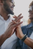 Two individuals are shown smiling and holding hands intimately. The focus is on their interlocked fingers, conveying a sense of connection and affection. One person is wearing a watch and a bracelet, while both are wearing casual clothing.
