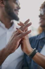 Two individuals are shown smiling and holding hands intimately. The focus is on their interlocked fingers, conveying a sense of connection and affection. One person is wearing a watch and a bracelet, while both are wearing casual clothing.