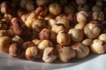 A close-up view of a pile of hazelnuts resting on a surface. The lighting highlights the rich, brown shells and creamy textures of the nuts, creating a warm and natural look.