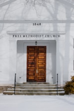 An old church building with a symmetrical facade featuring large wooden double doors and a small porch. The building is white with the number 1848 prominently displayed above the entrance. The words 'Free Methodist Church' are written in capital letters under the cornice. Bare tree branches are visible in the upper part of the image, casting shadows on the building.