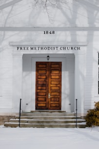 An old church building with a symmetrical facade featuring large wooden double doors and a small porch. The building is white with the number 1848 prominently displayed above the entrance. The words 'Free Methodist Church' are written in capital letters under the cornice. Bare tree branches are visible in the upper part of the image, casting shadows on the building.