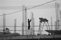 A person stands on a ladder working on a construction site with metal rods and a concrete structure. The scene includes scaffolding, wires, and construction equipment against a gray, overcast sky. The overall ambiance is industrial and a bit stark, captured in black and white.