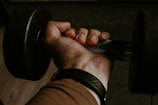 A strong hand gripping a dumbbell with Maschio Suplementos bottle in the background.