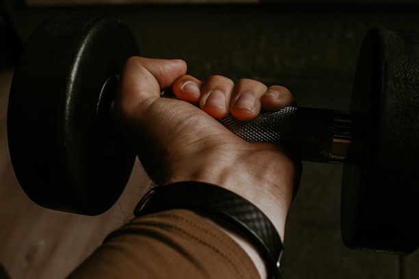 A close-up view of a hand gripping a dumbbell, showcasing the knurled texture of the handle. The skin appears slightly tanned, and the veins on the hand are subtly visible. The person is wearing a black wristband and a brown sleeve.