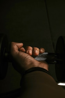 A close-up view of a hand gripping a metal dumbbell in dim lighting. The texture of the dumbbell handle and the details of the fingers are clearly visible. The shadowing and lighting create a focused and intense atmosphere.