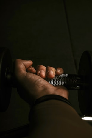 A close-up view of a hand gripping a metal dumbbell in dim lighting. The texture of the dumbbell handle and the details of the fingers are clearly visible. The shadowing and lighting create a focused and intense atmosphere.