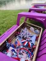 A wicker basket filled with patriotic-themed items, including ribbons and decorations in red, white, and blue with star patterns. The basket is placed on a purple outdoor chair with grass and a body of water in the background.