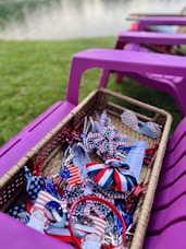 A wicker basket filled with patriotic-themed items, including ribbons and decorations in red, white, and blue with star patterns. The basket is placed on a purple outdoor chair with grass and a body of water in the background.